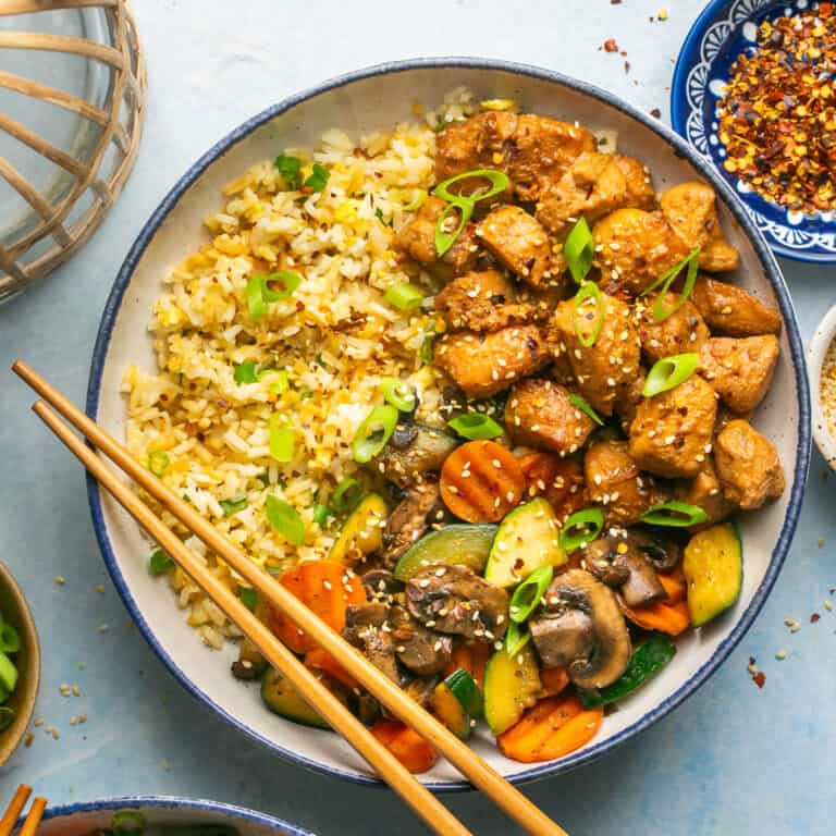 A bowl of marinated and grilled chicken over fried rice with hibachi style vegetables and chop sticks.