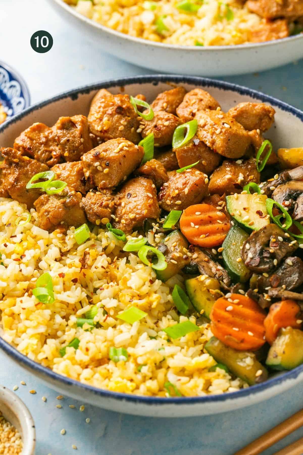 Close up image of the assembled hibachi bowls with marinated cooked chicken breast, fried rice and sauteed vegetables with green onions and sesame seeds on top. 