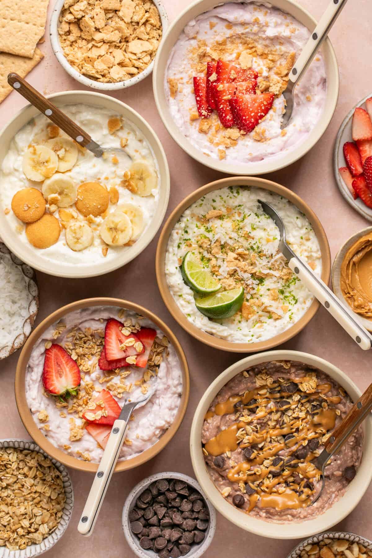 Very Berry, Key Lime Pie, Banana Cream Pie, Strawberry Cheesecake and Chocolate Peanut Butter cottage cheese bowls laid out with extra toppings around and spoons for serving. 