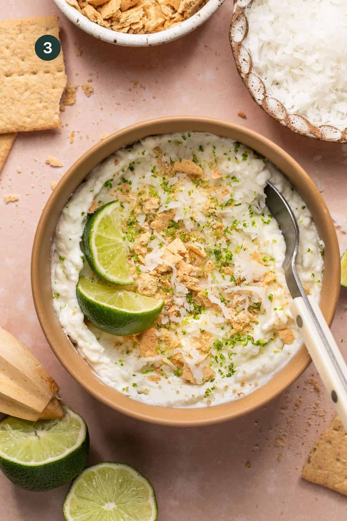 Coconut flakes, graham cracker crumbs and lime zest added to the top of a cottage cheese bowl with a spoon and toppings sprinkled around. 