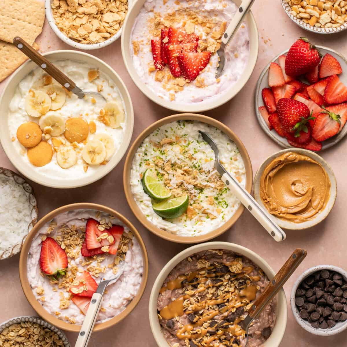 Very Berry, Key Lime Pie, Banana Cream Pie, Strawberry Cheesecake and Chocolate Peanut Butter cottage cheese bowls laid out with extra toppings around and spoons for serving.