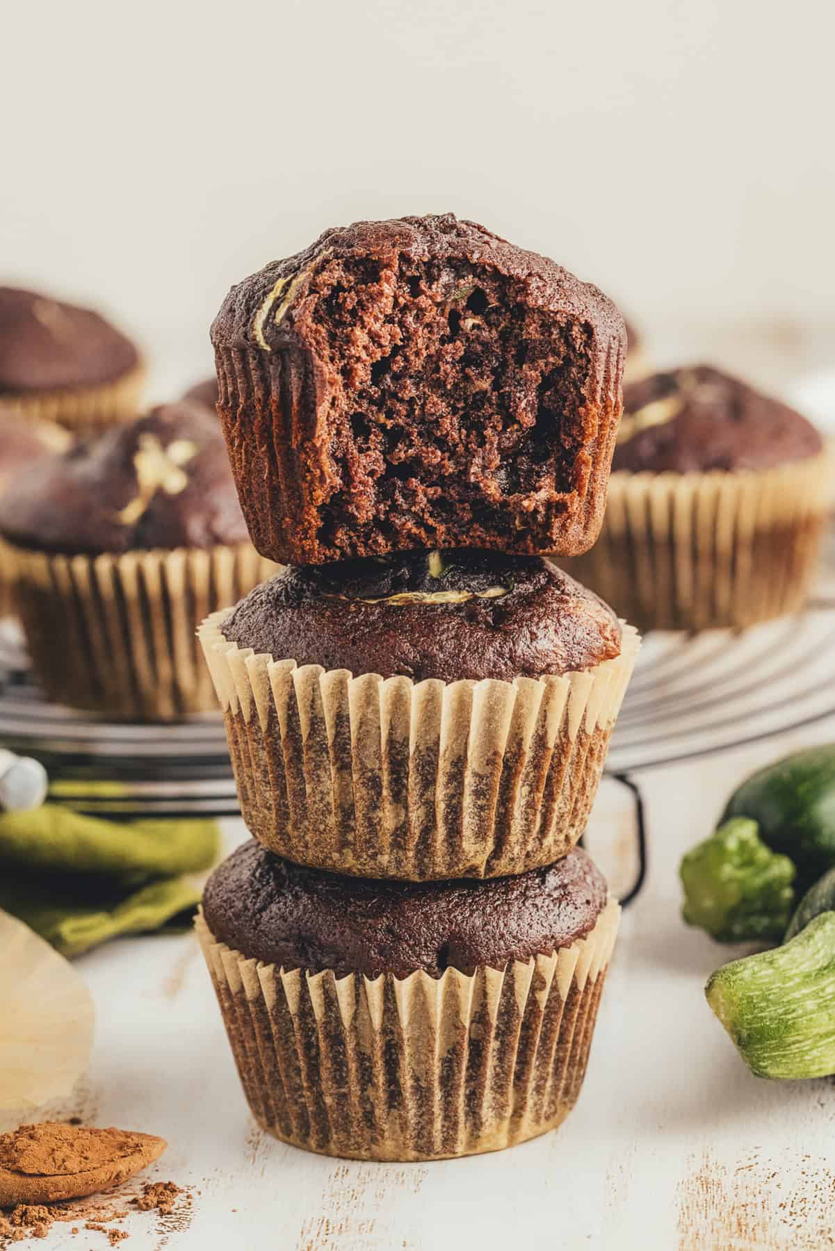 Three stacked zucchini chocolate muffins with 1 with a bite out of it and other muffins arranged behind it. 