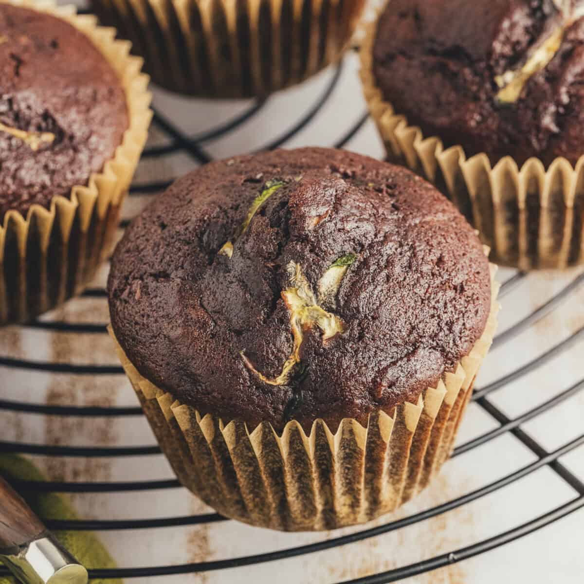 One chocolate zucchini muffin in a muffin wrapper on a cooling rack with three muffins arranged behind it.