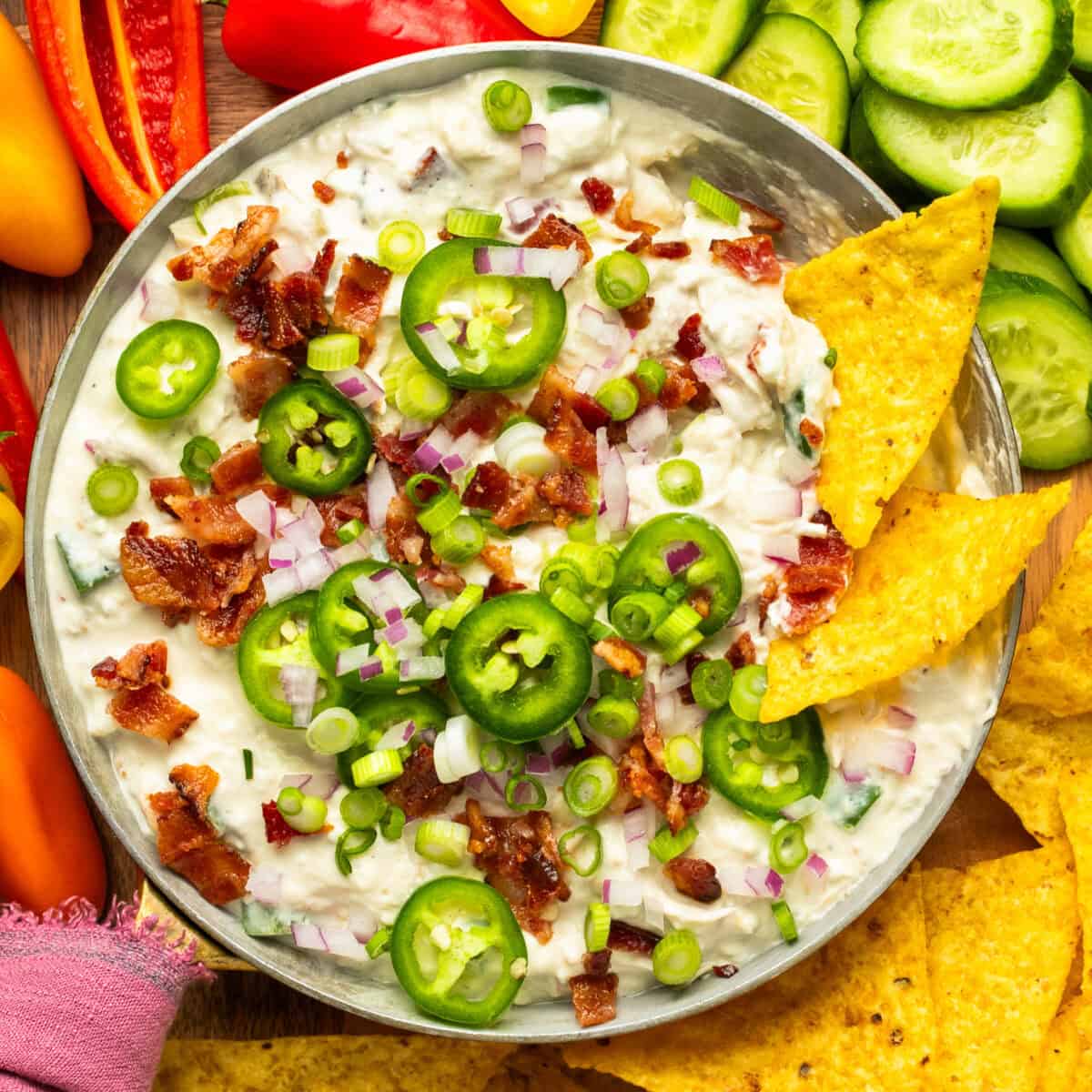 Jalapeno popper dip in a serving bowl topped with bacon, onions, jalapenos and chips and veggies around for serving.