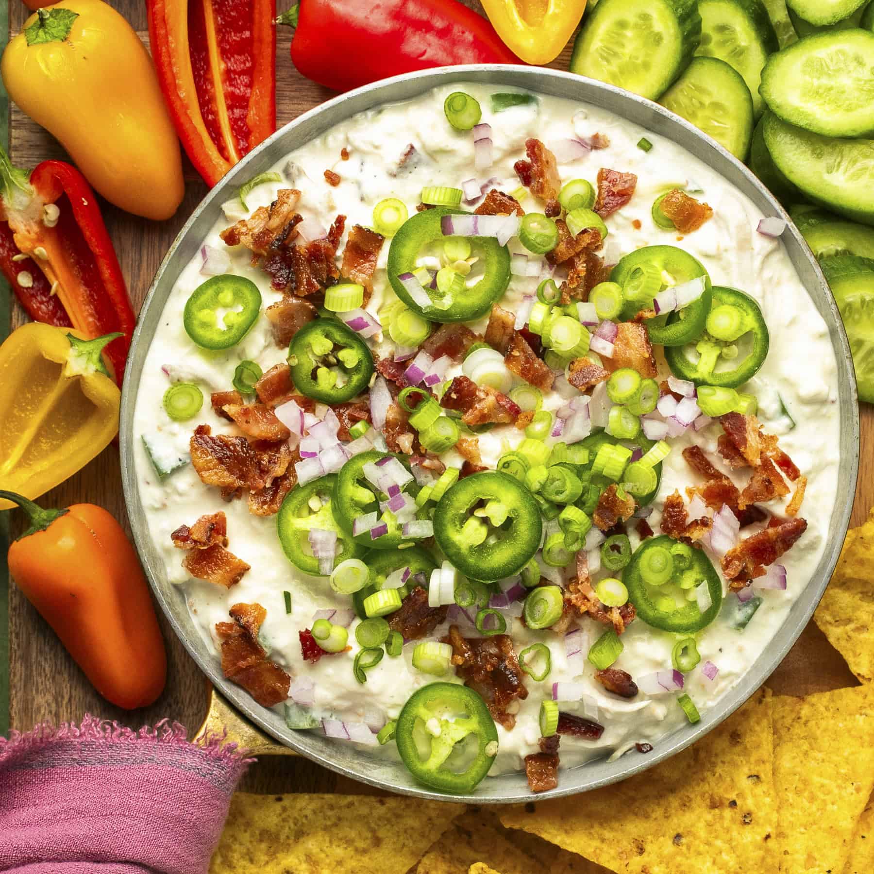 Jalapeno popper dip in a serving bowl with jalapenos, bacon and onions on top with veggies and chips around for dipping.
