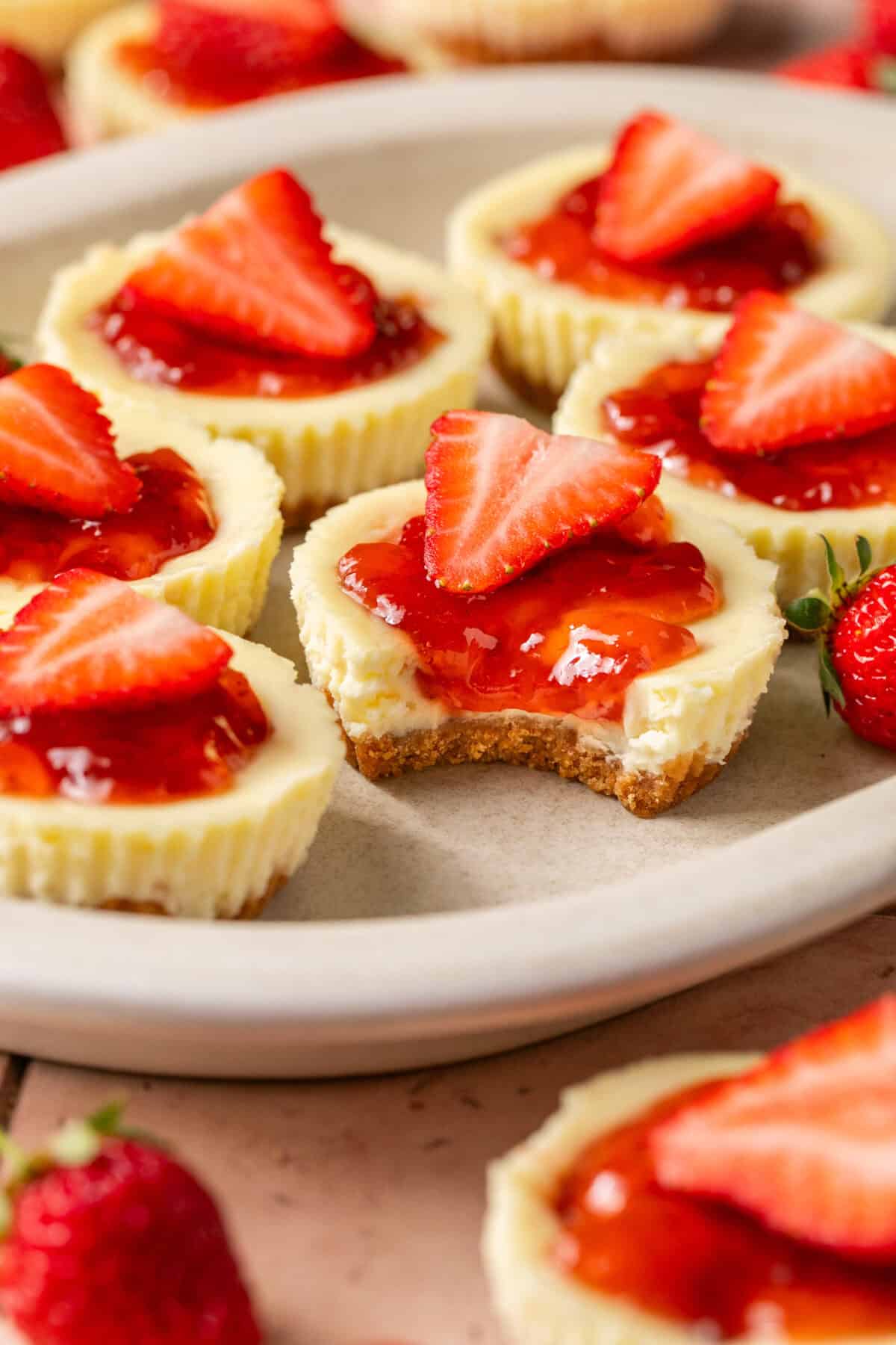 Mini cheesecakes laid out on a plate with strawberry jam on top and fresh strawberries and a bite taken out of one. 