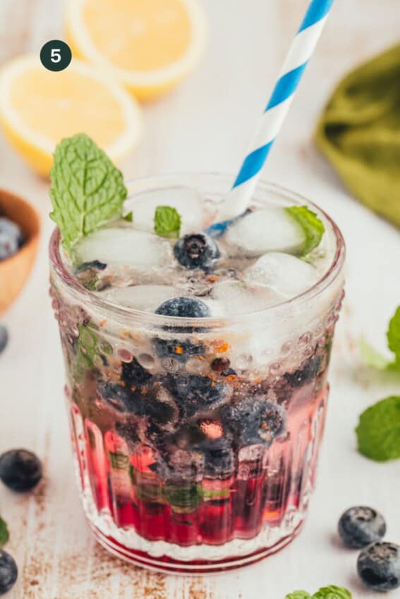 Blueberries in a glass with ice and a muddled honey juice with coconut water on top and a straw garnished with mint.