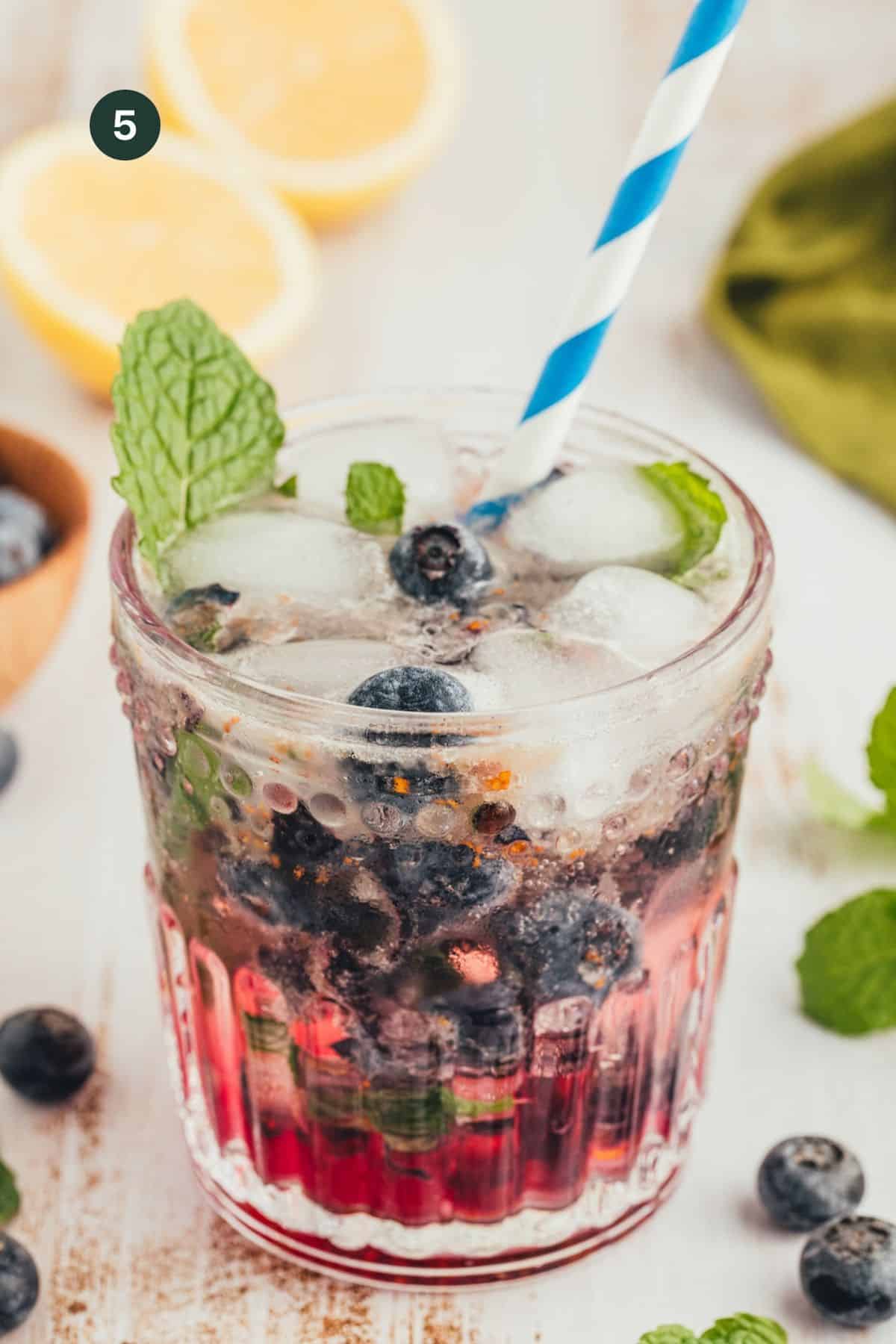 Blueberries in a glass with ice and a muddled honey juice with coconut water on top and a straw garnished with mint. 