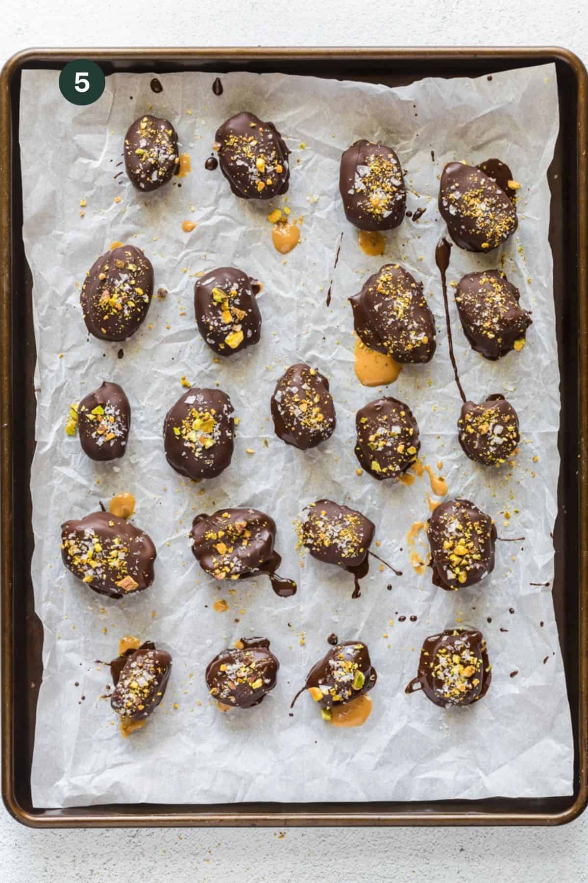 Baking sheet full of chocolate covered dates with salt and pistachios crumbled on top. 
