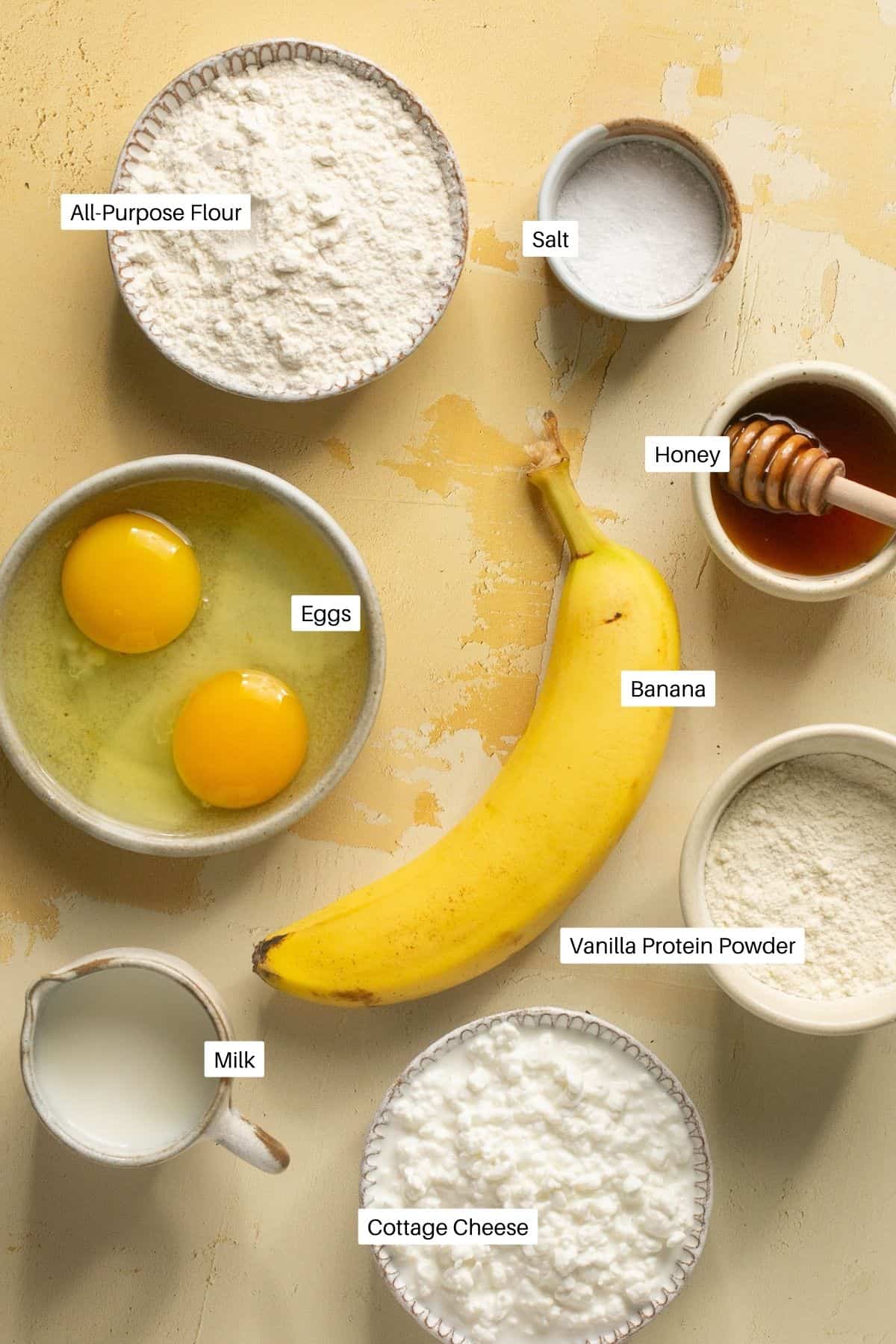 Flour, salt, honey, banana, eggs, milk, cottage cheese and protein powder laid out to make waffles. 