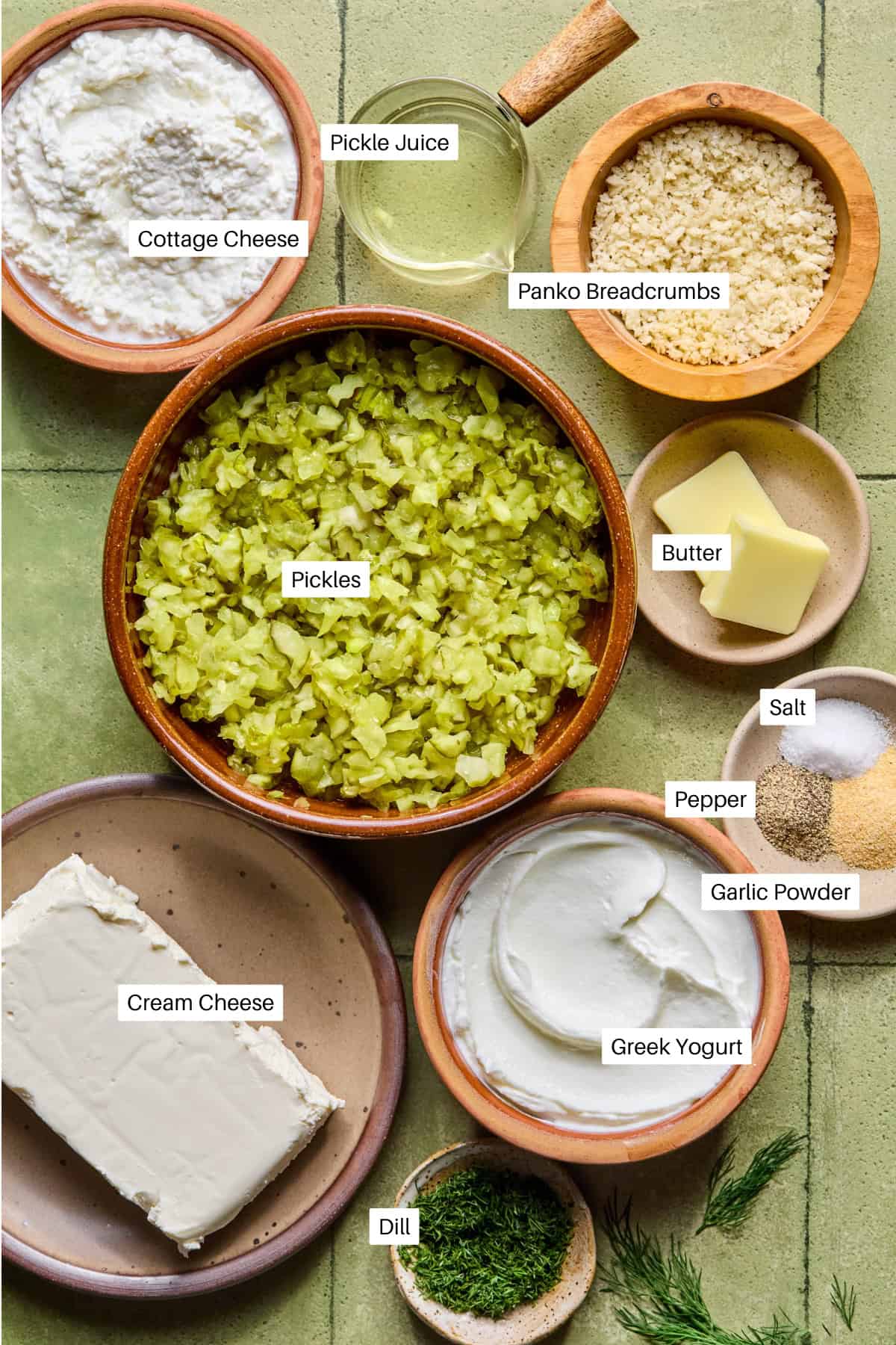 A top-down view of labeled ingredients for fried pickle dip in bowls and plates: cottage cheese, pickle juice, panko breadcrumbs, butter, pickles, salt, pepper, garlic powder, Greek yogurt, cream cheese, and dill on a green tile surface.