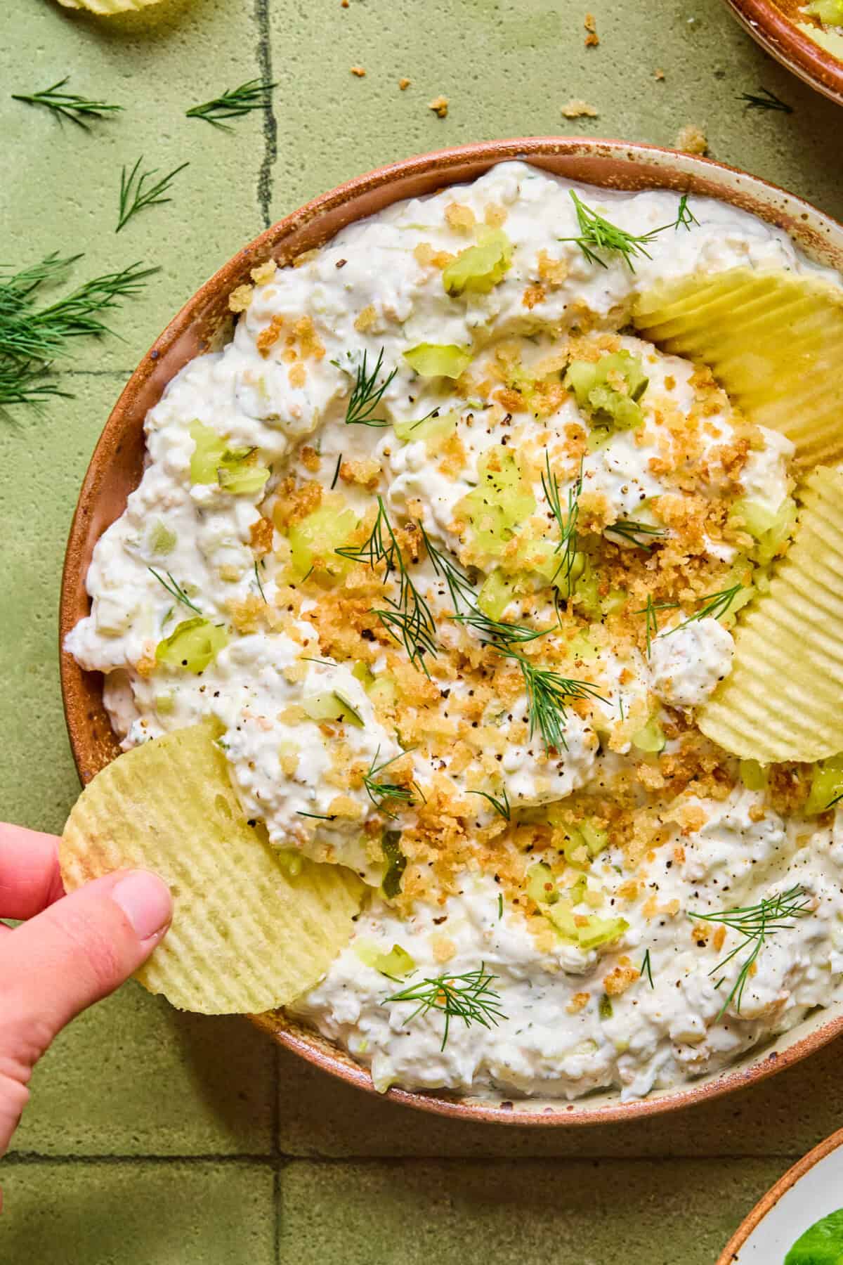 A hand dips a ridged potato chip into a bowl of creamy fried pickle dip, garnished with fresh dill, chopped celery, black pepper, and crunchy crumbs, set on a green tiled surface.