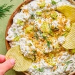 A hand dips a ridged potato chip into a bowl of creamy fried pickle dip, garnished with fresh dill, chopped celery, black pepper, and crunchy crumbs, set on a green tiled surface.
