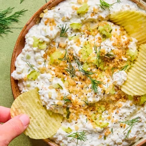 A hand dips a ridged potato chip into a bowl of creamy fried pickle dip, garnished with fresh dill, chopped celery, black pepper, and crunchy crumbs, set on a green tiled surface.