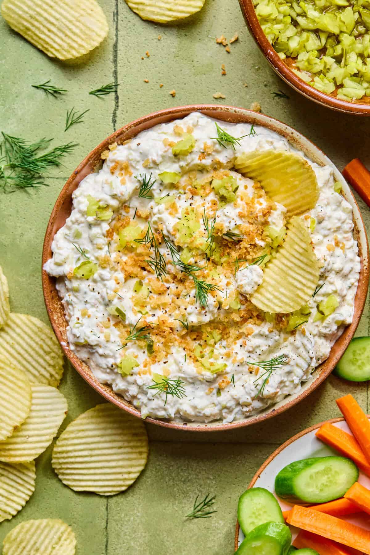 A bowl of creamy fried pickle dip garnished with crushed potato chips, chopped celery, and fresh dill, surrounded by ridged potato chips and a plate of sliced cucumbers and carrot sticks.