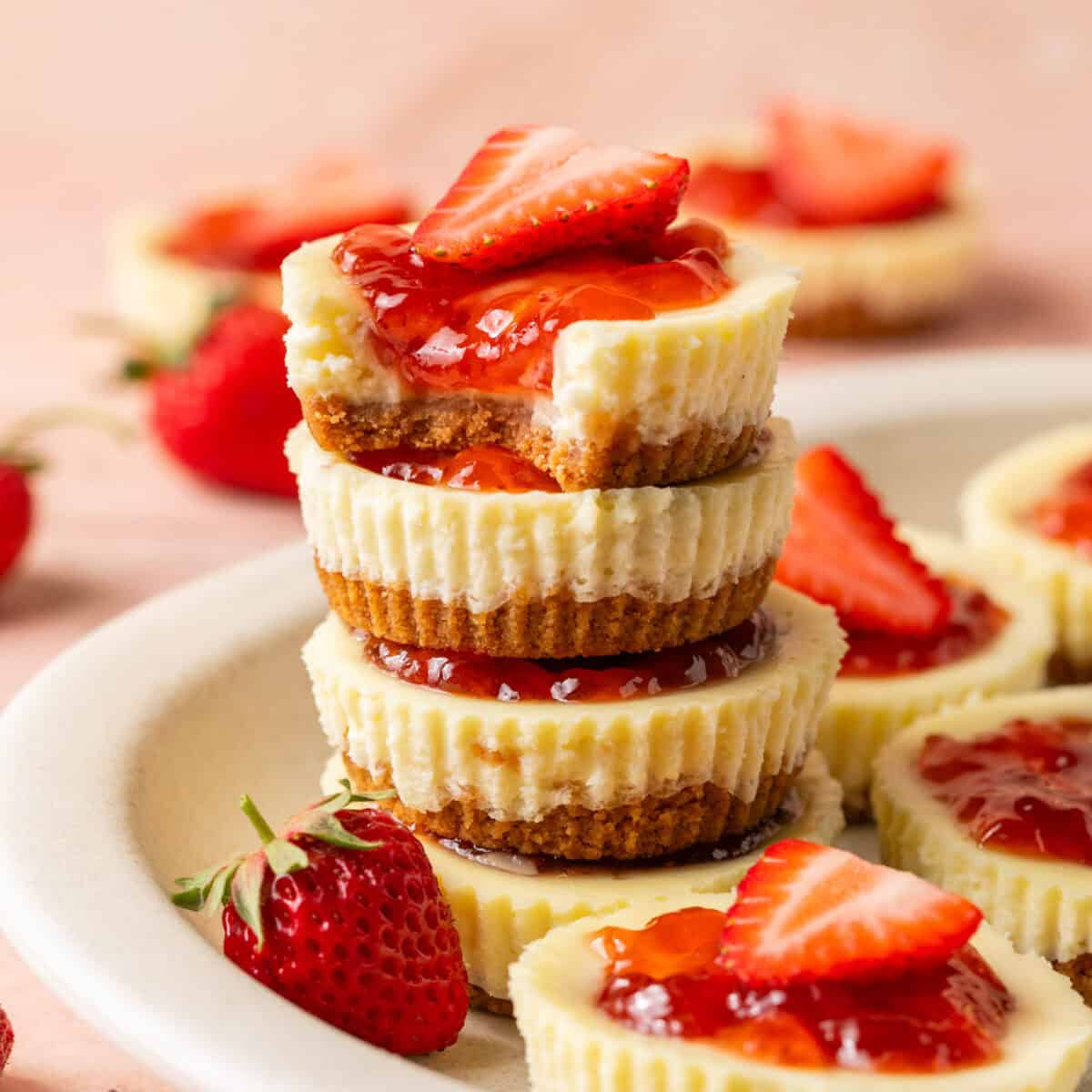 4 stacked mini cheesecakes with a bite taken out of the top one and strawberry jam on top and fresh strawberries on a plate.