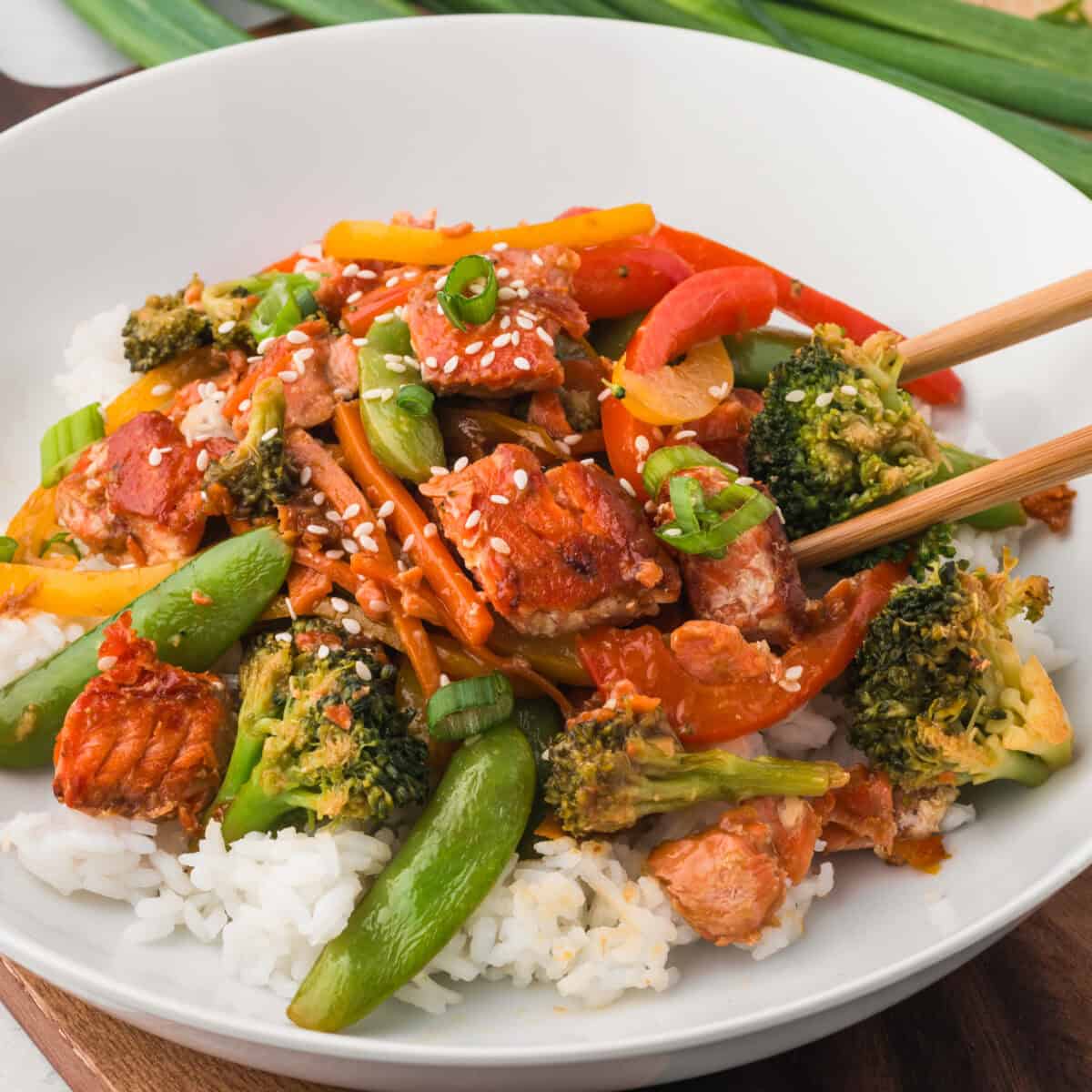 Salmon stir fry with stir fry veggies over rice on a place with chop sticks.