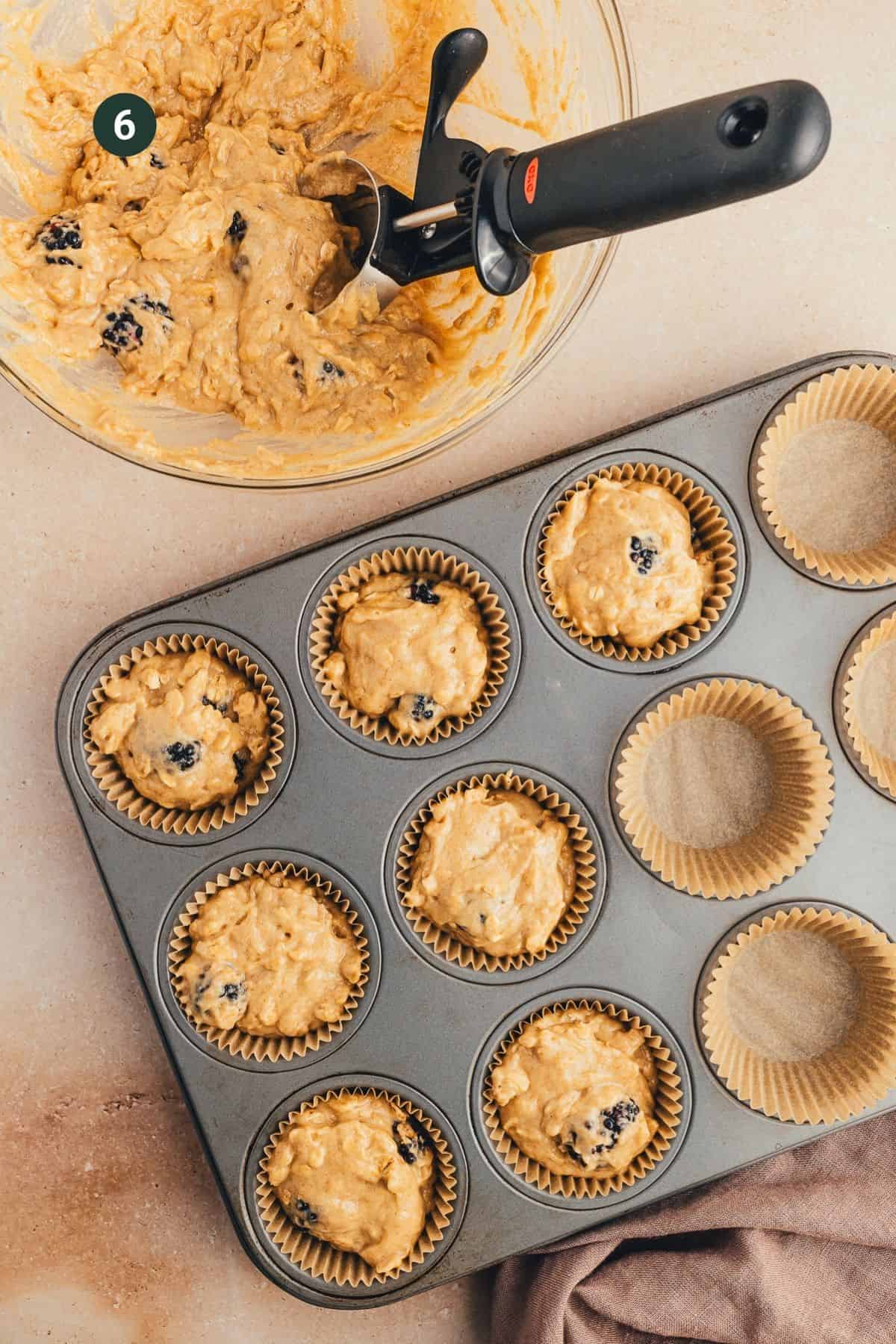 Blackberry muffin batter placed in lined muffin cups with a scooper.