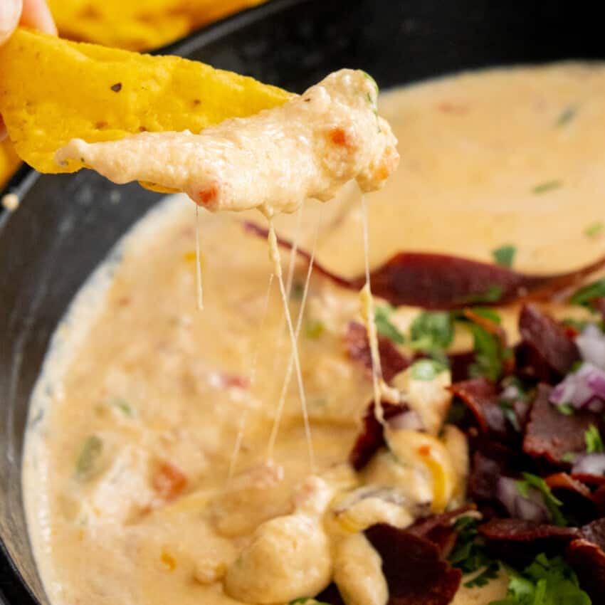 A cheesy nacho being dipped into a creamy queso dip garnished with chopped herbs, red onions, and slices of meat. The cheese stretches between the nacho and the dip, reminiscent of decadent cottage cheese recipes that highlight its gooey texture.