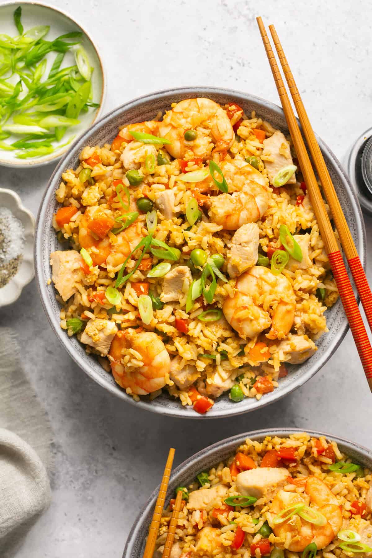 Chicken and shrimp stir fry in a bowl with chop sticks laying to enjoy with a small dish near with sliced green onions.