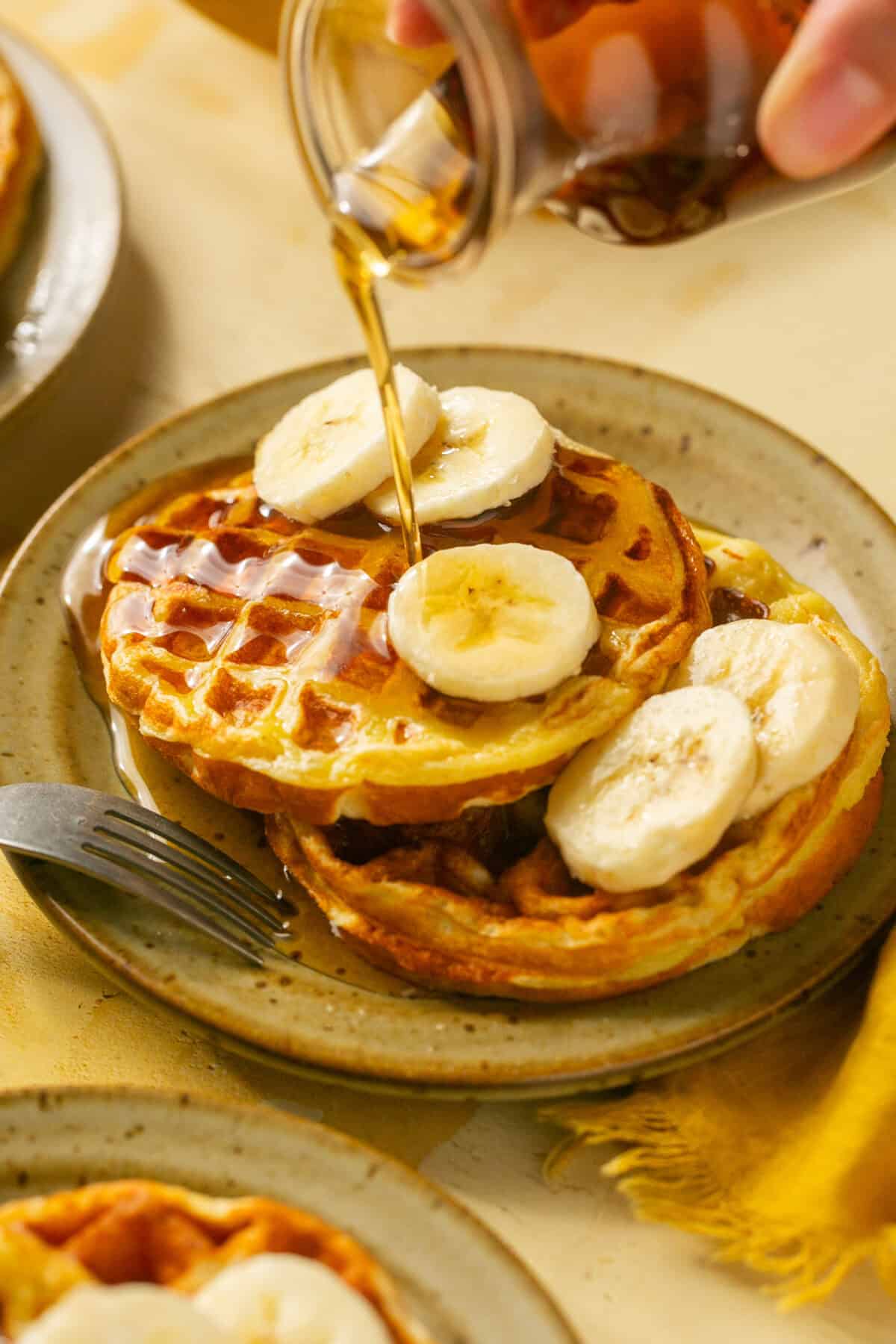 Two waffles on a plate with sliced banana, syrup being poured on top with a fork.