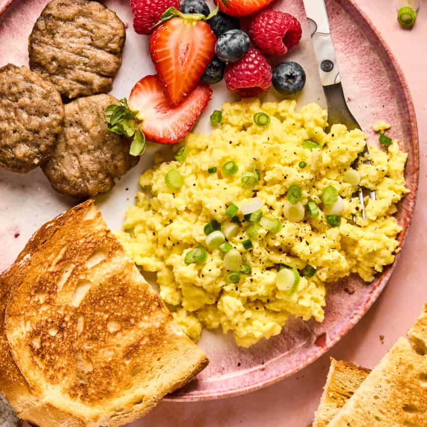 A pink plate features scrambled eggs topped with green onions, fresh berries, sausage patties, and toasted bread slices. Perfect for incorporating into your cottage cheese recipes, a fork rests on the side to complete this delightful breakfast ensemble.