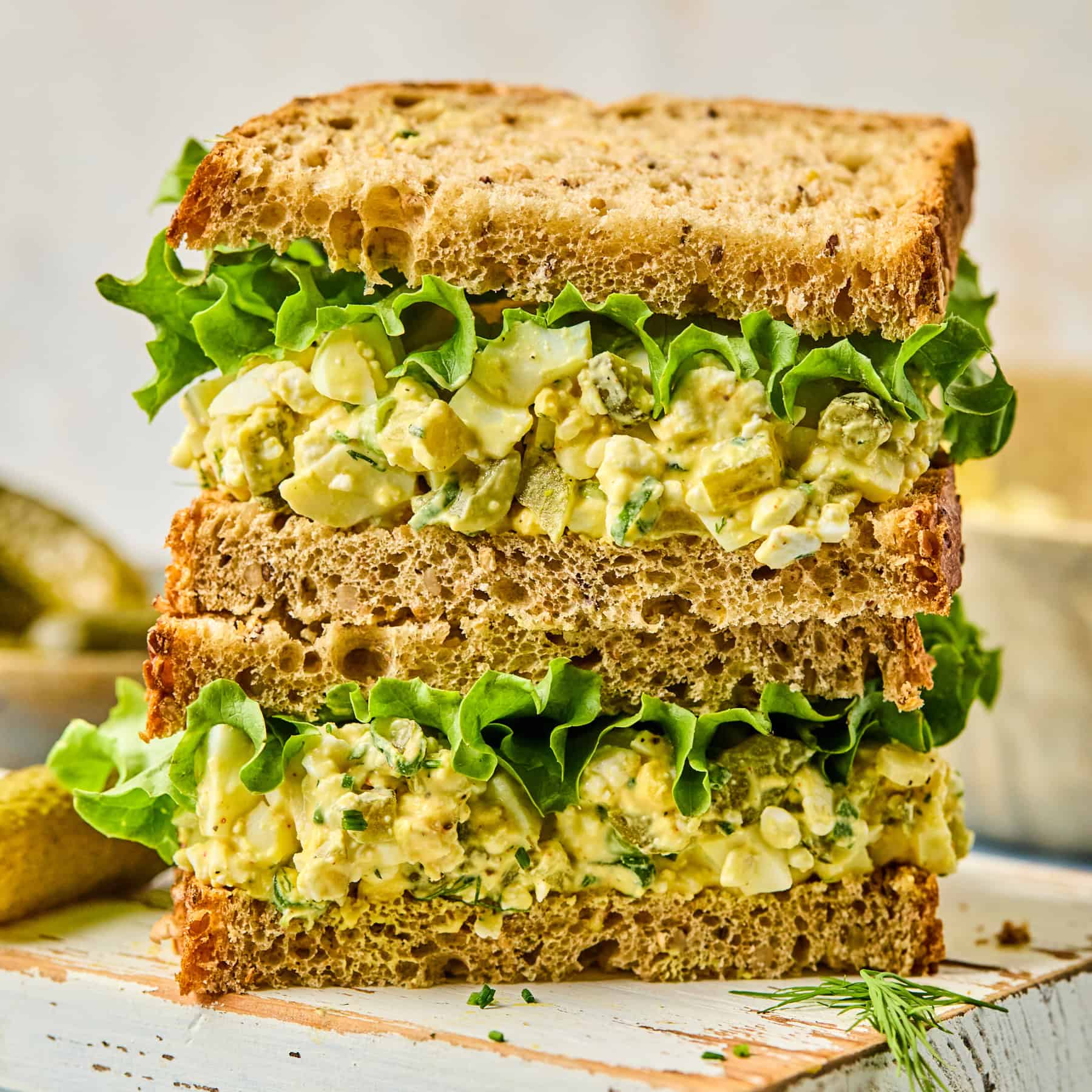 Close-up of a sandwich with two layers, filled with egg salad and crisp green lettuce between slices of multigrain bread, set on a rustic wooden surface reminiscent of cottage cheese recipes. A few pickles are blurred in the background.