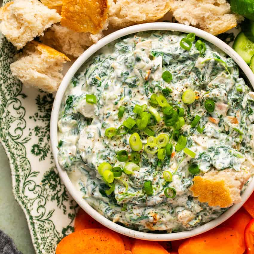 Spinach dip in a serving dish topped with green onions and a piece of crusty bread dipped in surrounded by more bread cubes and carrot slices.