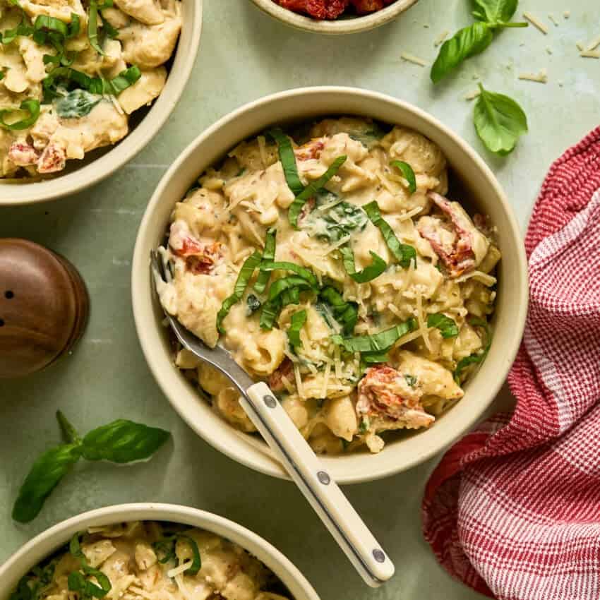 A bowl of creamy pasta, reminiscent of delightful cottage cheese recipes, is garnished with sliced basil leaves and shredded cheese. The dish includes chunks of sun-dried tomatoes. A fork rests on the bowls edge amid fresh basil, a salt shaker, and a red checkered cloth.