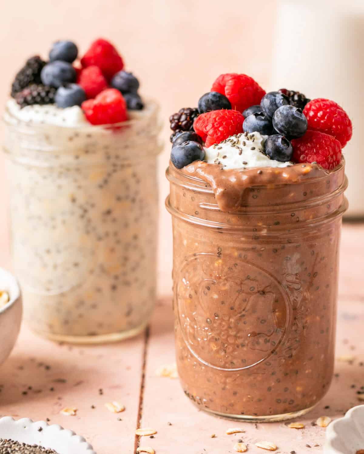 Two jars of overnight oats one chocolate and the other vanilla both topped with mixed berries, greek yogurt and sprinkle of chia seeds.