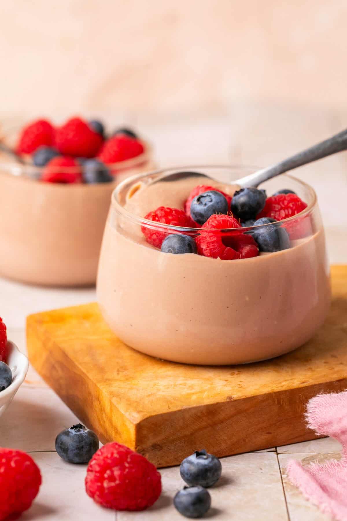 Pudding in a pudding cup with a spoon and berries on top and one behind it. 