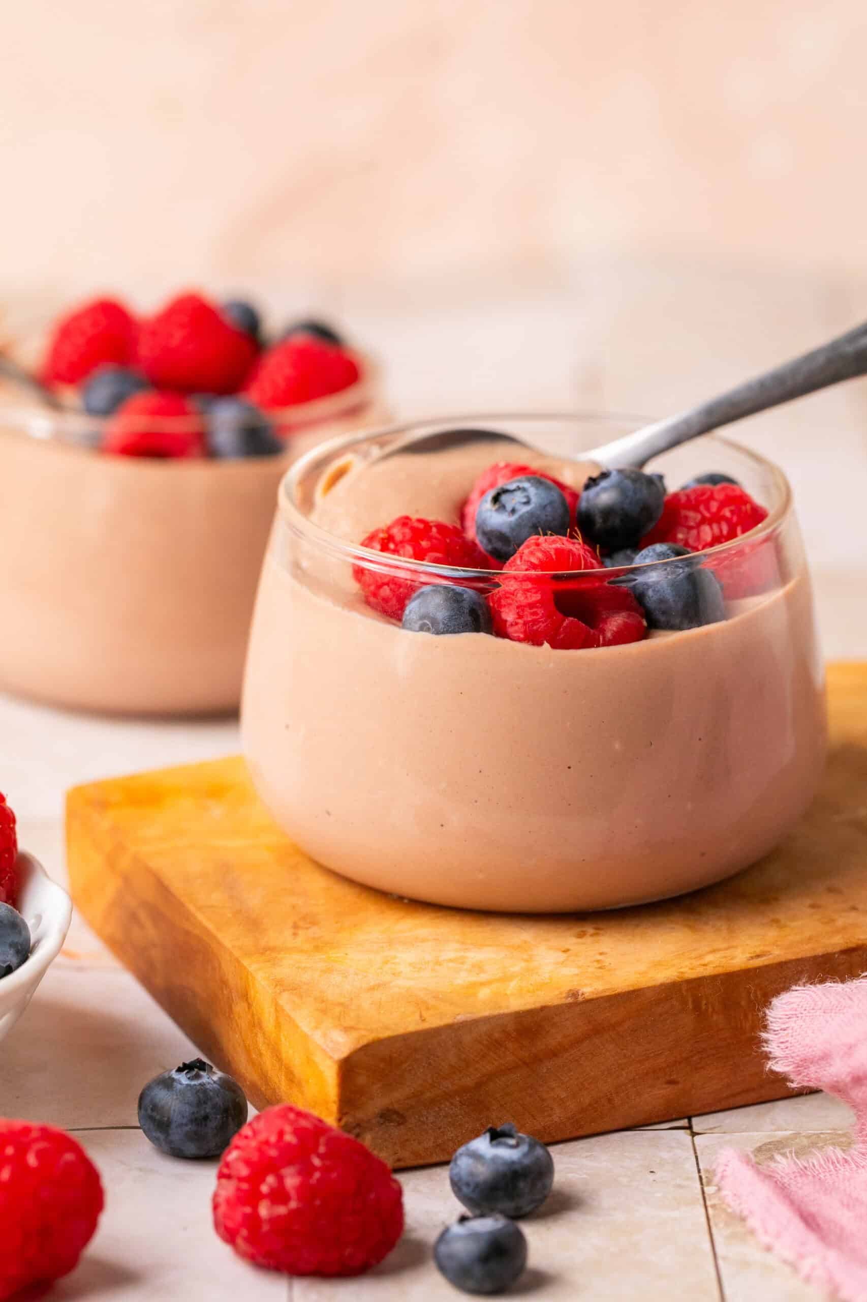 Pudding in a pudding cup with a spoon and berries on top and one behind it.