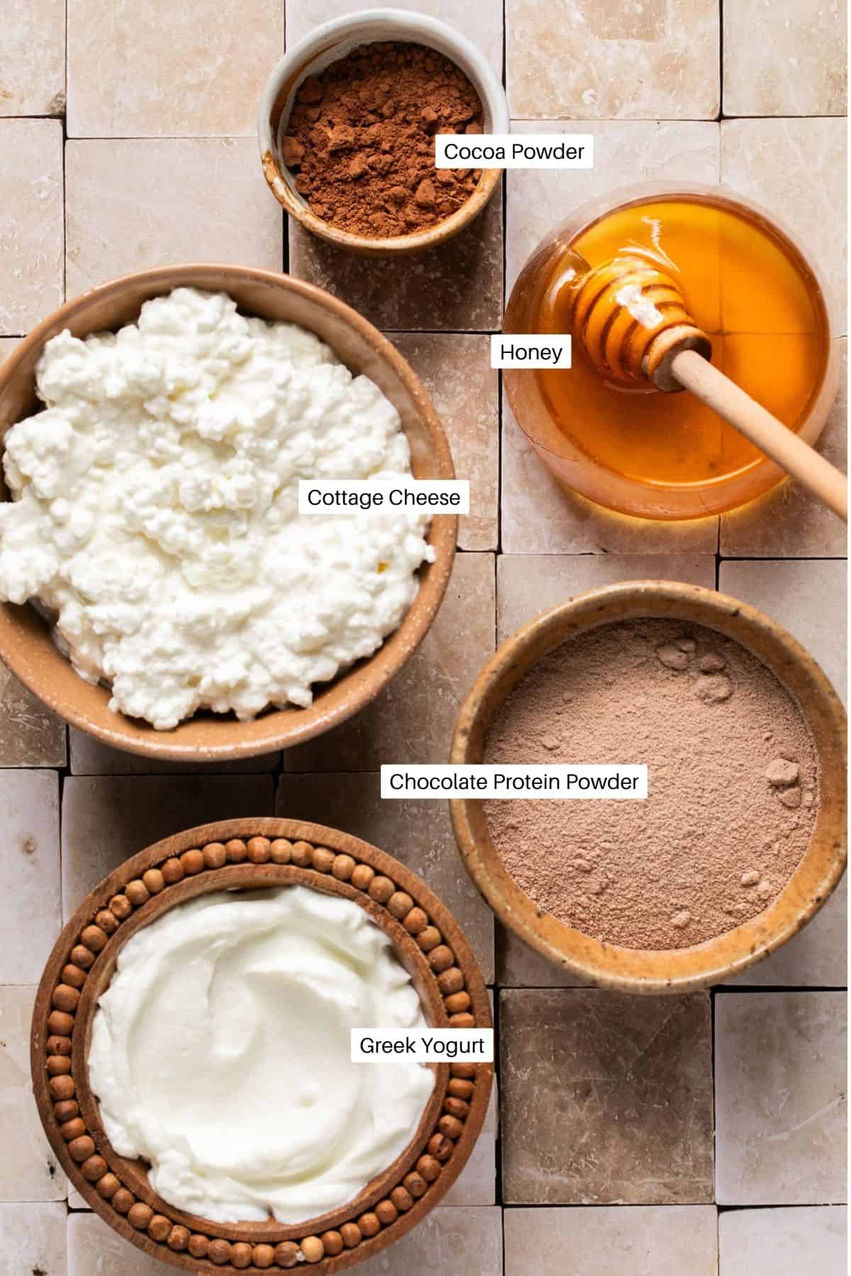 Cocoa powder, honey, cottage cheese, greek yogurt and protein powder laid out to make pudding. 