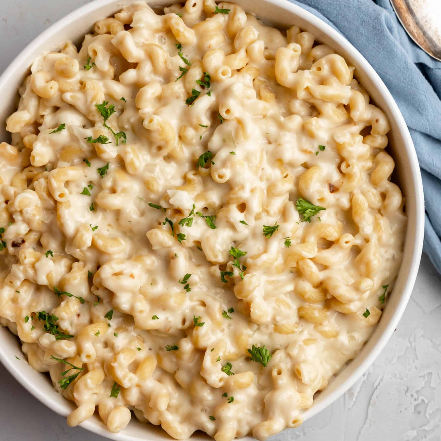 A bowl of creamy macaroni and cheese, inspired by classic cottage cheese recipes, is garnished with chopped parsley. It rests on a light-colored surface near a blue napkin and a spoon.