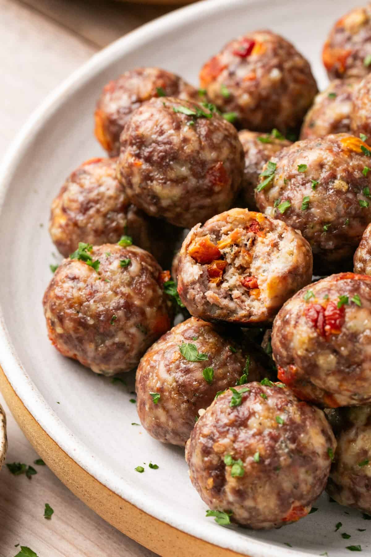 Close up of a meatball with a bite taken out of it, surrounded by a plate of cooked meatballs. 