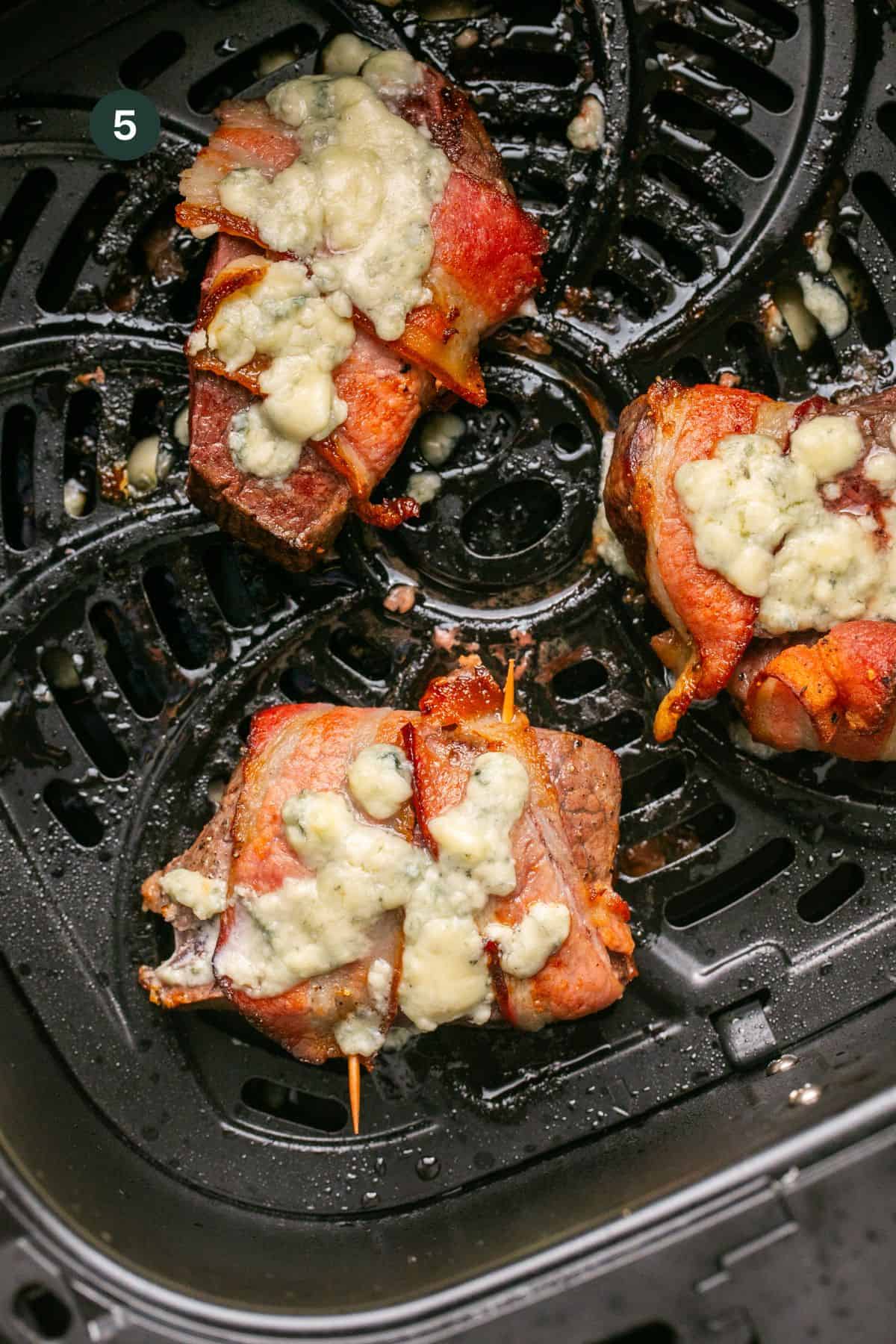 Filet mignon in the air fryer basket wrapped in bacon, cooked and topped with melted gorgonzola cheese. 