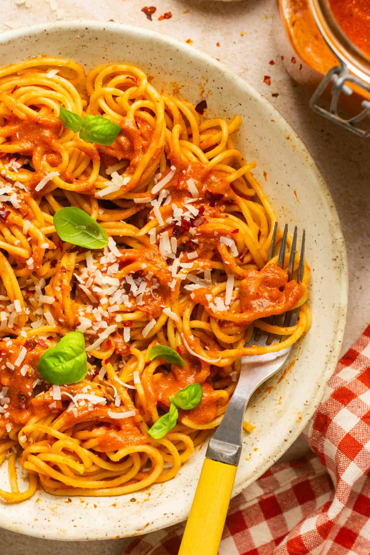 Spaghetti noodles with pasta sauce, grated parmesan cheese and fresh basil on a place to serve with a red and white gingham towel and fork. 