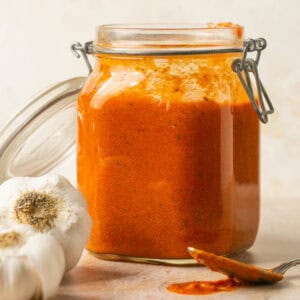 Creamy tomato sauce in a sealable jar with the lid open and a spoon with sauce on it laid out and garlic on the side.