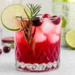 Cranberry lime drink in a rocks glass with ice and garnished with rosemary spring, cranberries and lime.