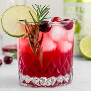 Cranberry lime drink in a rocks glass with ice and garnished with rosemary spring, cranberries and lime.