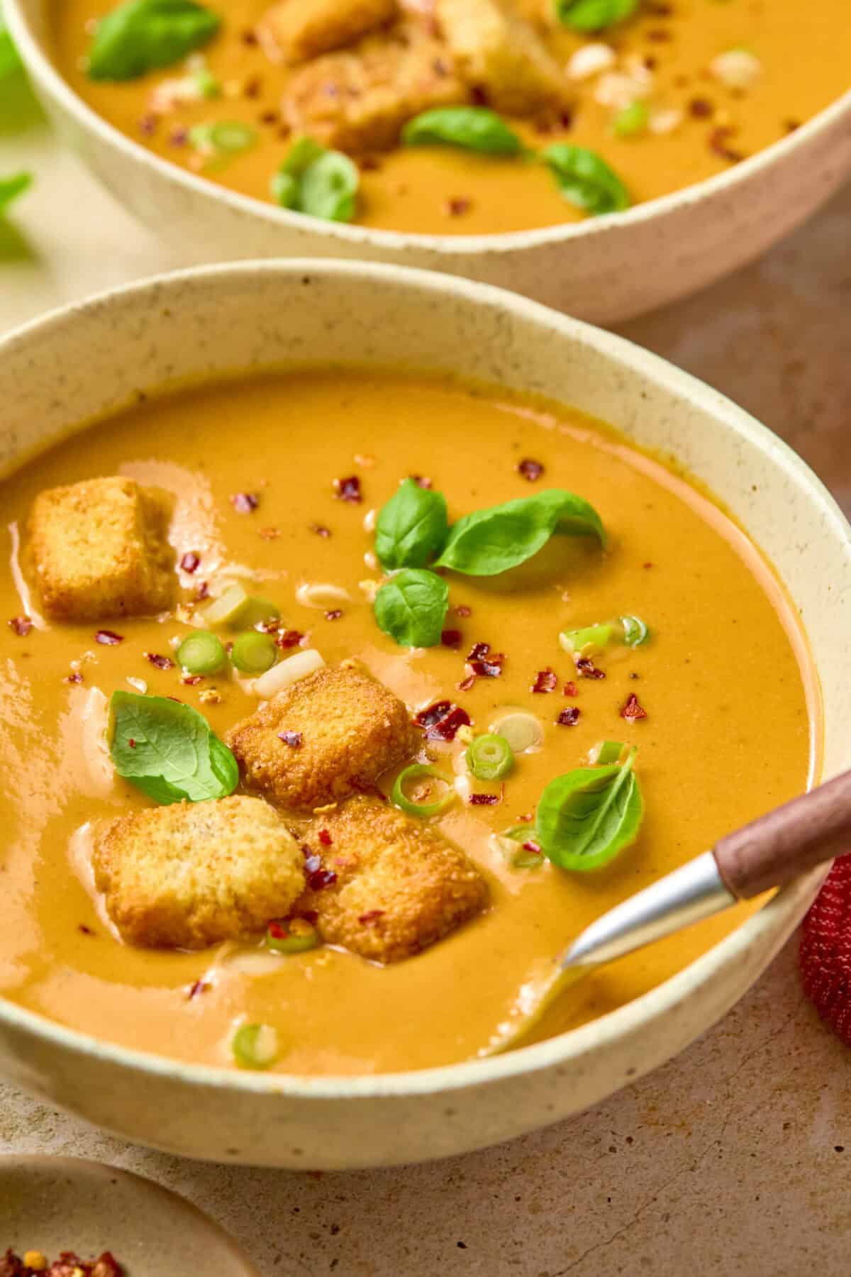 A bowl of butternut squash soup topped with basil, green onions, red pepper flakes and croutons with a spoon taking a spoonful out to enjoy.