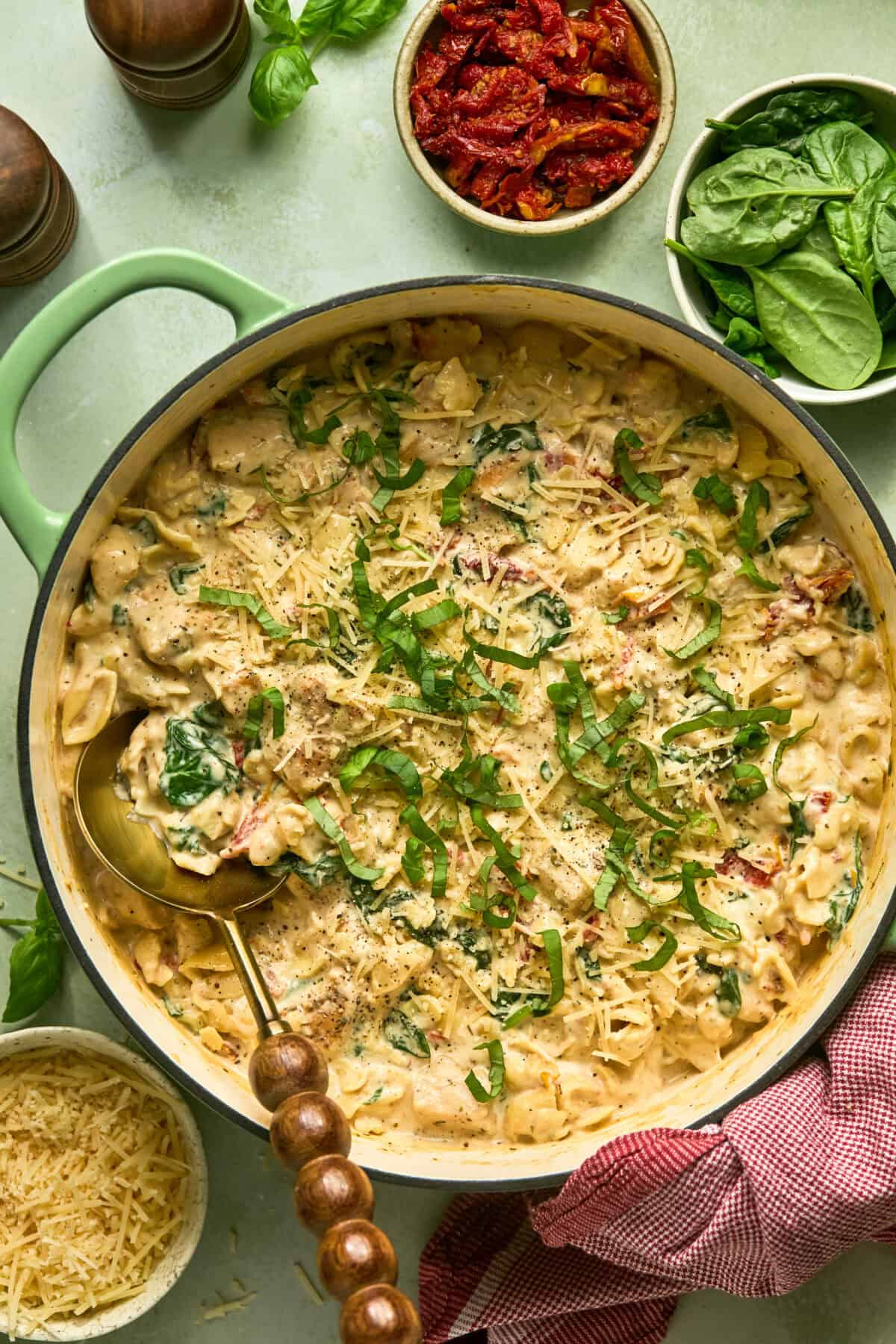 A large skillet full of creamy pasta with sun dried tomatoes and basil strips on top and spoon for serving.