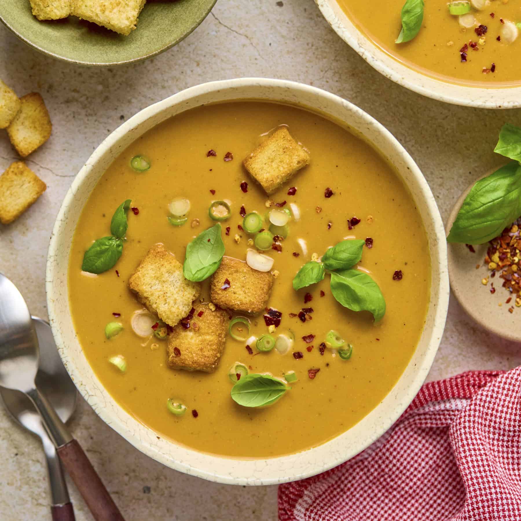 A bowl of soup topped with basil, green onions, red pepper flakes and croutons with a spoon for serving.