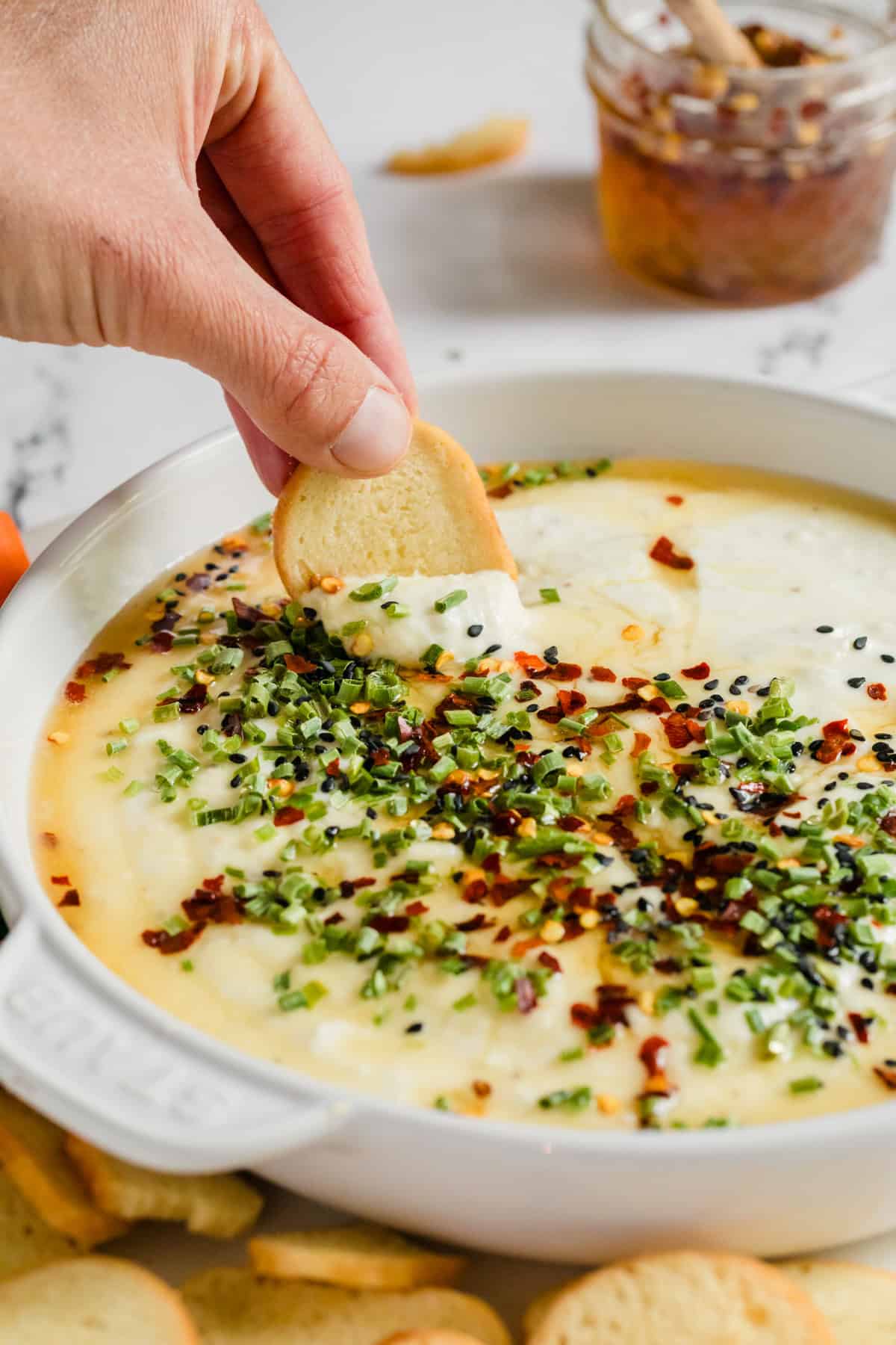 Bowl of creamy dip with chives and honey on top and a hand scooping some out with a cracker.