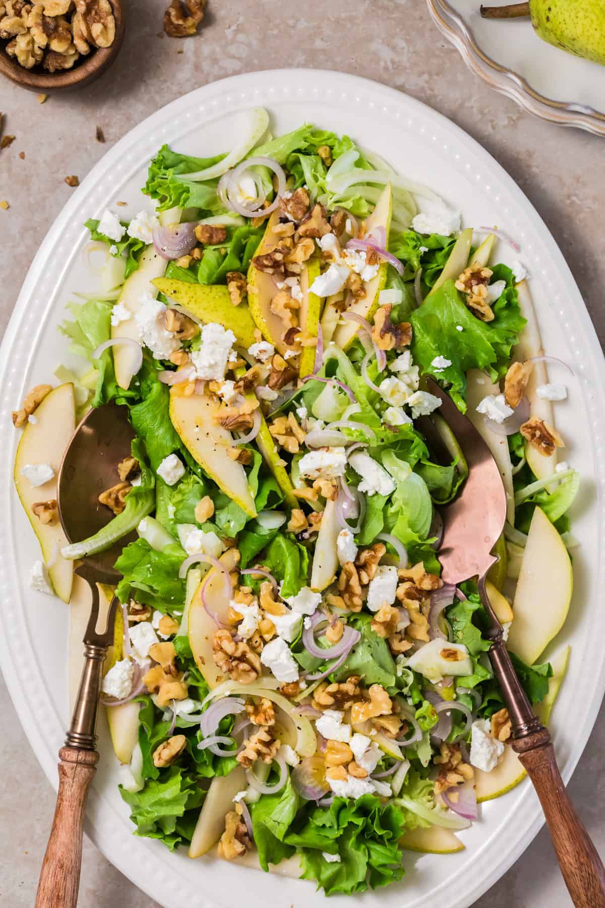 Mixed greens, pears and feta salad with serving utensils on a dish. 