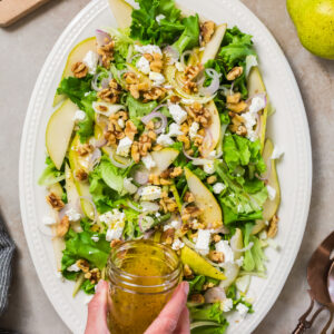 Salad with walnuts, feta and sliced pears on a serving dish with dressing being poured on top.