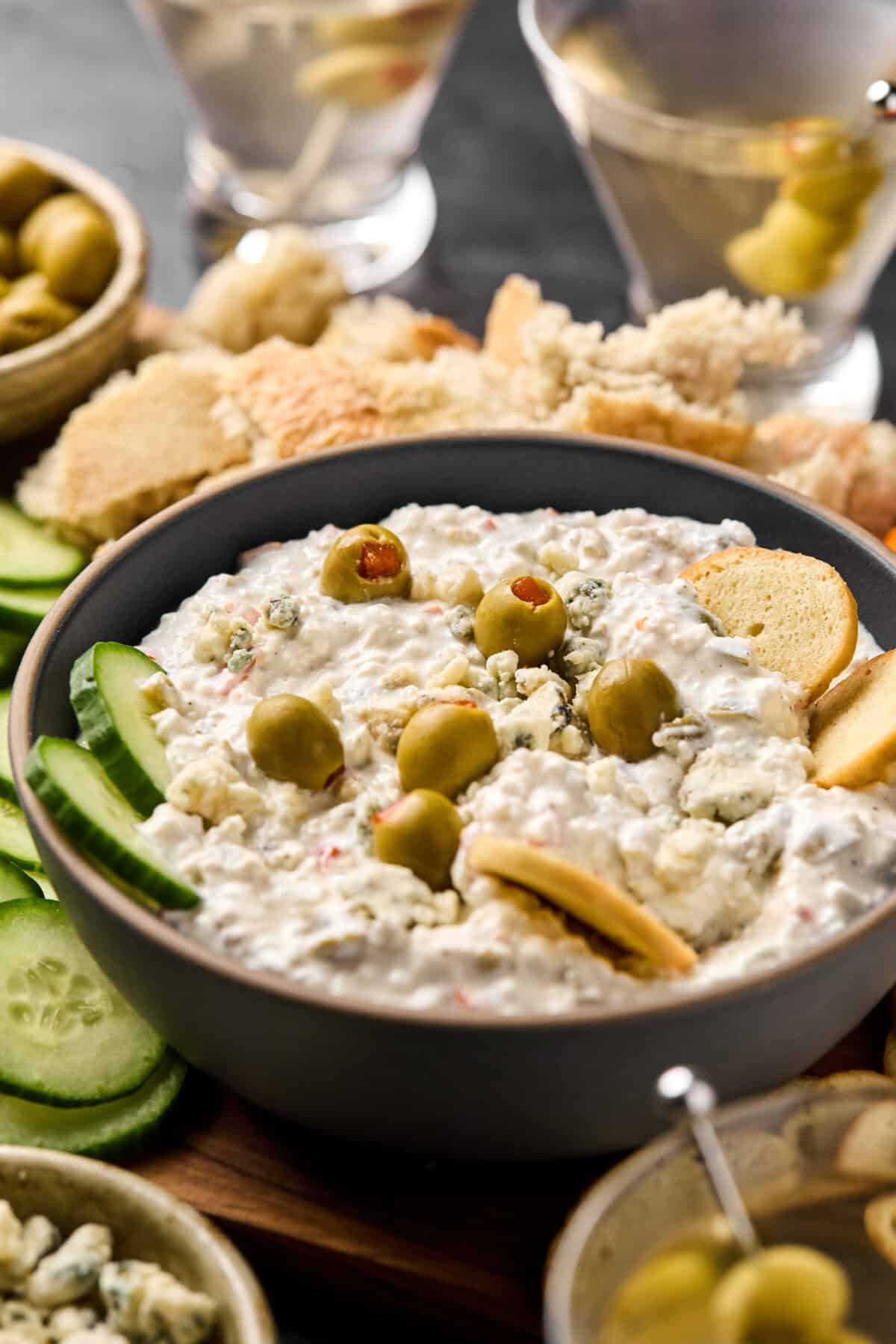 A bowl of creamy dirty martini dip garnished with green olives. Surrounding the bowl are slices of cucumber, pieces of bread, and more olives. In the background, two elegant martini glasses complete the scene.