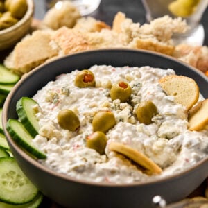 A bowl of creamy dirty martini dip garnished with green olives. Surrounding the bowl are slices of cucumber, pieces of bread, and more olives. In the background, two elegant martini glasses complete the scene.