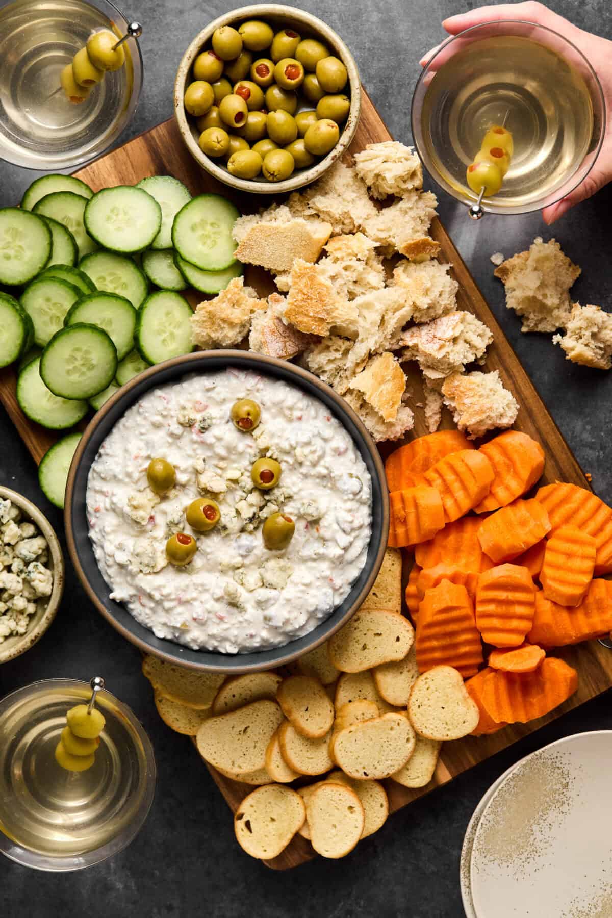 A wooden board showcases a bowl of creamy, dirty martini dip topped with olives, surrounded by sliced cucumbers, carrots, baguette pieces, and crackers. Glasses of martinis with olives are on the side, one being held by a hand.