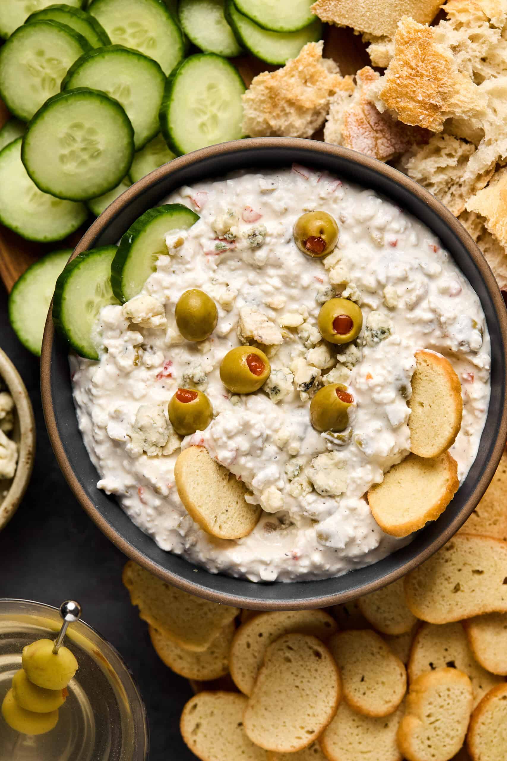 A bowl of creamy dirty martini dip topped with green olives is surrounded by cucumber slices, torn bread, and round toasted crackers. A small dish containing toothpicked olives sits beside the bowl, making for an elegant appetizer spread.